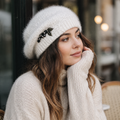 Woman wearing a white knit hat with a decorative brooch and scarf, sitting outdoors.