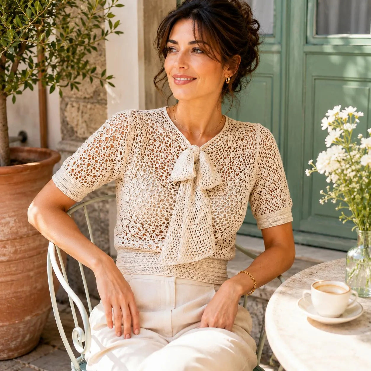 A woman sits outdoors at a table wearing a cream crochet short-sleeve top with a large bow at the neckline and cream pants. The setting includes a potted tree, a green door, and a small table with a cup of coffee and white flowers.