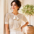 A woman wearing a cream-colored crochet short-sleeve top with a bow detail at the neckline, standing in a sunlit indoor setting with a potted plant in the background.