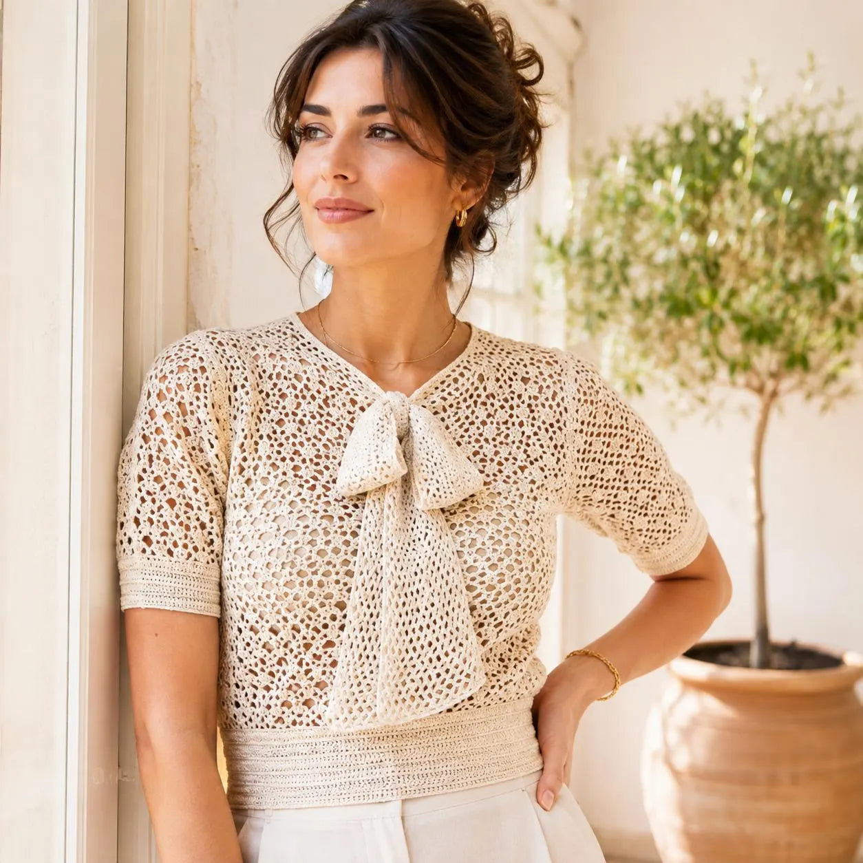A woman wearing a cream-colored crochet short-sleeve top with a bow detail at the neckline, standing in a sunlit indoor setting with a potted plant in the background.