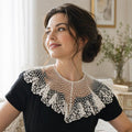 A woman wearing a black short-sleeve top with a large white crocheted ruffled collar, photographed indoors against a softly lit neutral background.