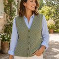 A woman wearing a sage green cable-knit button-front vest over a light blue button-up shirt and cream pants, standing outdoors in a garden courtyard.