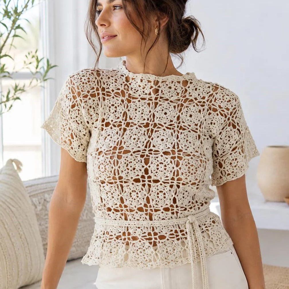 A woman wearing a cream crochet lace short-sleeve top with a ruffled hem and tie detail, paired with cream pants, standing in a bright, modern room.