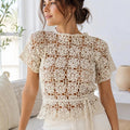 A woman wearing a cream crochet lace short-sleeve top with a ruffled hem and tie detail, paired with cream pants, standing in a bright, modern room.