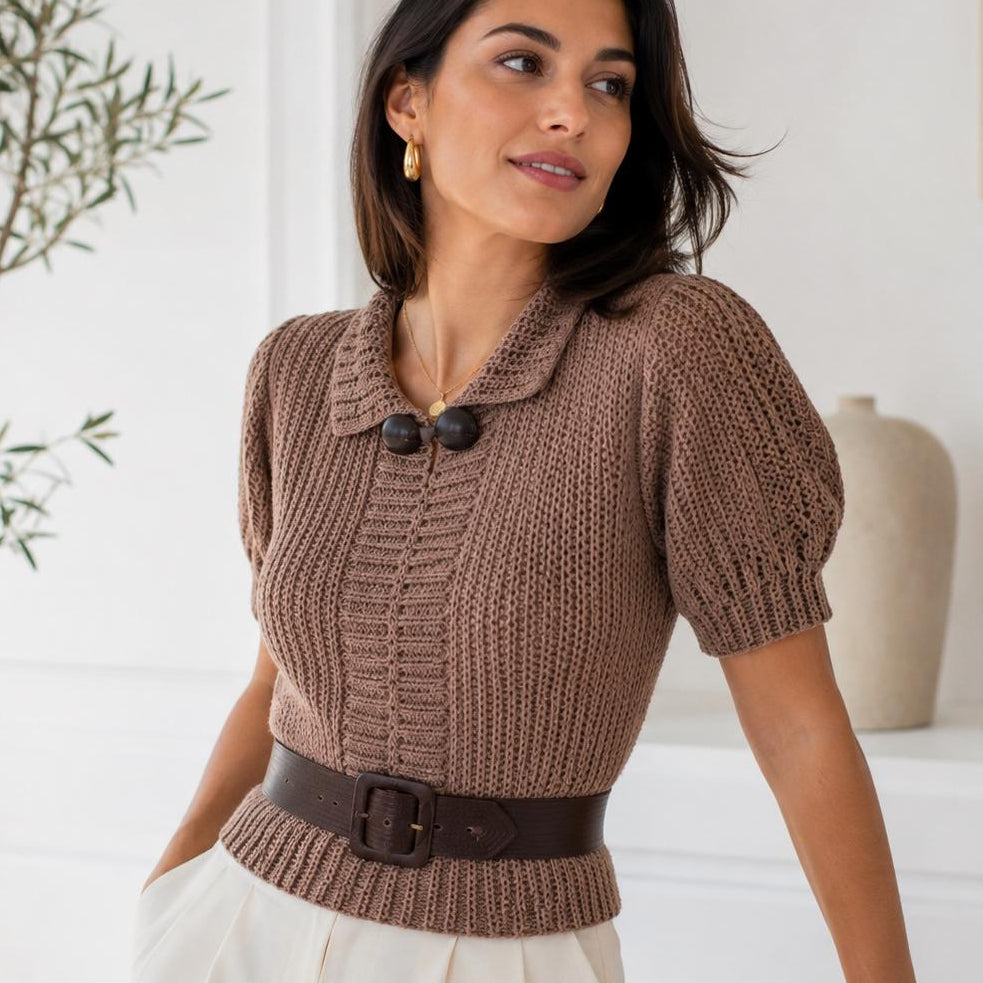 A woman wearing a brown chunky knit short-sleeve top with a collar and dark buttons, styled with a dark brown belt and white trousers, in a bright, modern room.