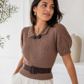 A woman wearing a brown chunky knit short-sleeve top with a collar and dark buttons, styled with a dark brown belt and white trousers, in a bright, modern room.