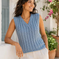 A woman wearing a sleeveless light blue cable-knit V-neck sweater vest, paired with white pants, leaning against a white wall in an outdoor setting with potted plants and pink flowers.