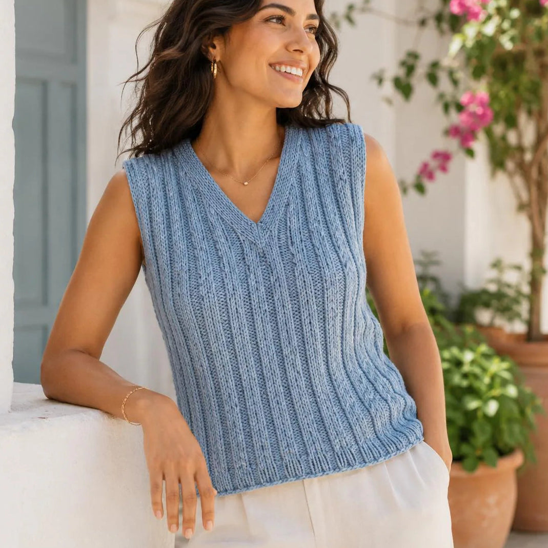 A woman wearing a sleeveless light blue cable-knit V-neck sweater vest, paired with white pants, leaning against a white wall in an outdoor setting with potted plants and pink flowers.
