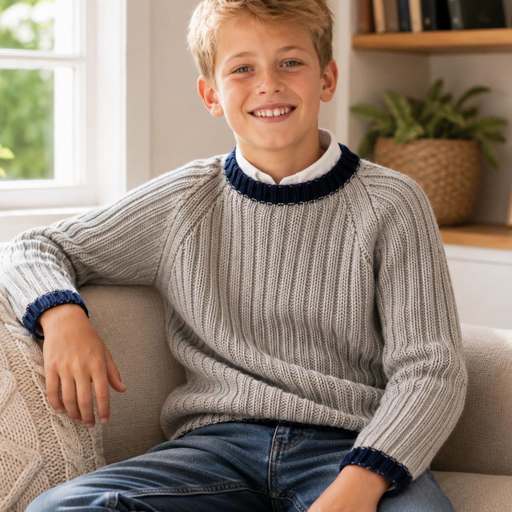 A smiling young boy sits on a beige couch wearing a ribbed knit crewneck sweater in light gray with dark navy trim, layered over a white collared shirt and paired with blue jeans.