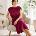 A woman wearing a textured deep burgundy midi dress sits in a light beige armchair in a bright, modern living room with potted plants and a side table.