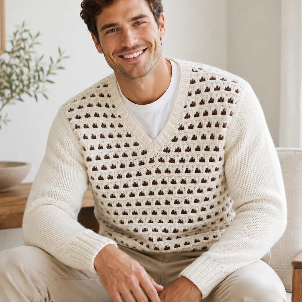 A smiling man seated in a light-colored armchair wearing a cream knit sweater vest with a repeating brown geometric pattern over a white T-shirt and beige pants, in a bright indoor room.