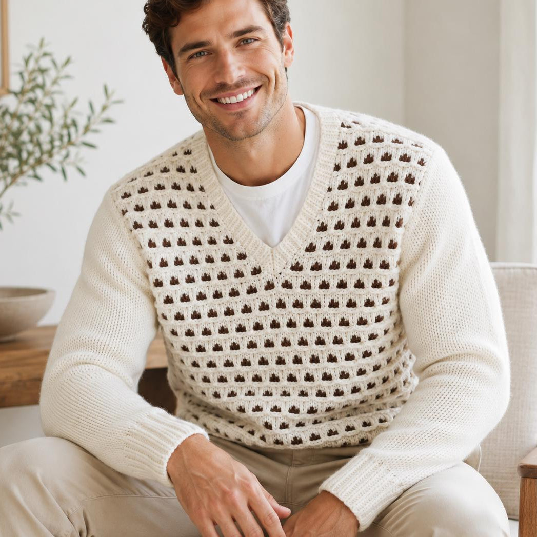 A smiling man seated in a light-colored armchair wearing a cream knit sweater vest with a repeating brown geometric pattern over a white T-shirt and beige pants, in a bright indoor room.